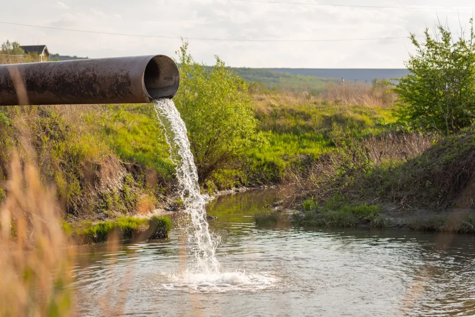 Large metal pipe discharging water or wastewater into creek or body of water with shrubs and field in background.